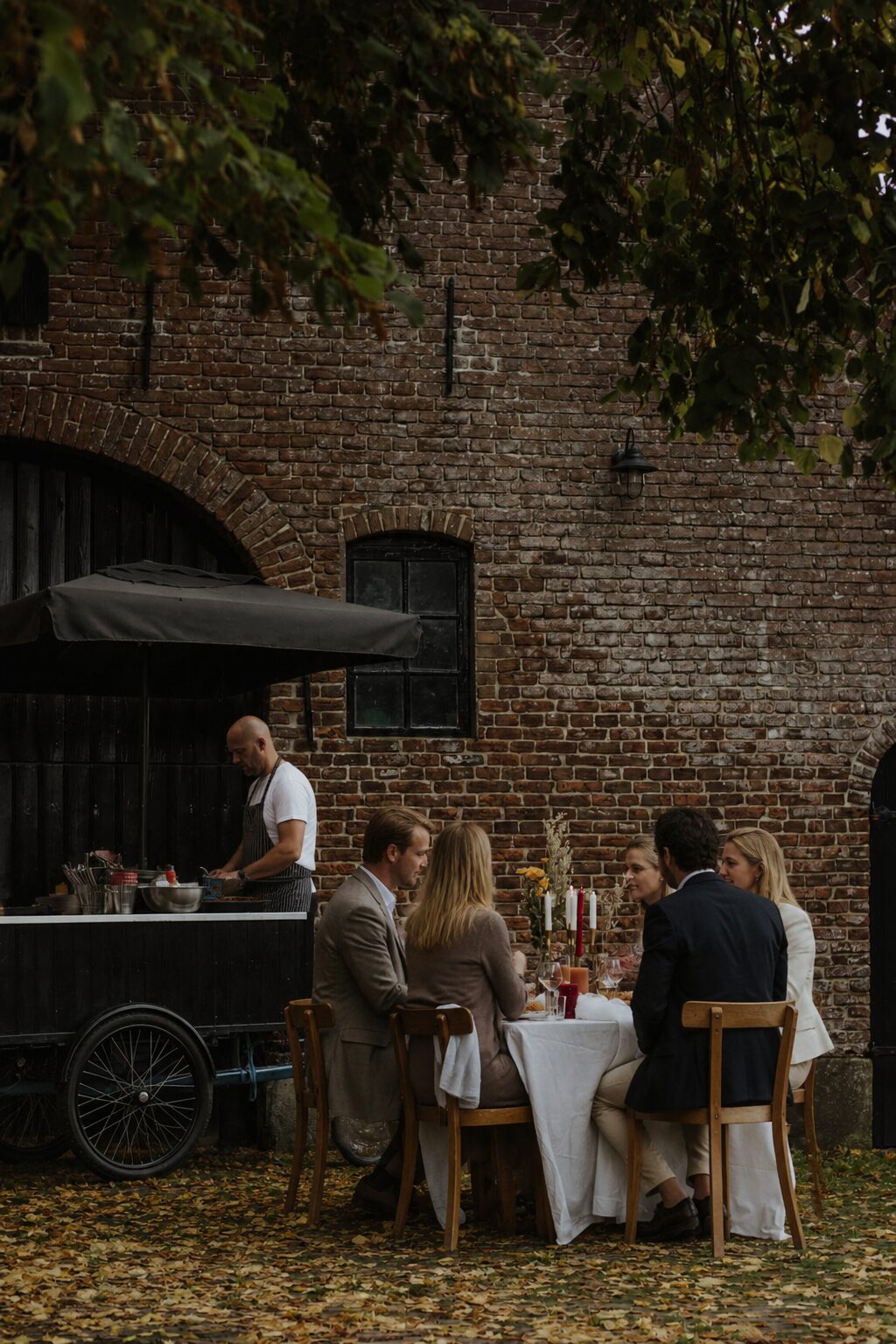 Kok bereidt gerechten bij de bakfiets terwijl gasten aan tafel zitten voor een oude schuur